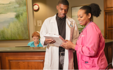 Two healthcare professionals, one in a white lab coat and the other in pink scrubs, discussing a clipboard.